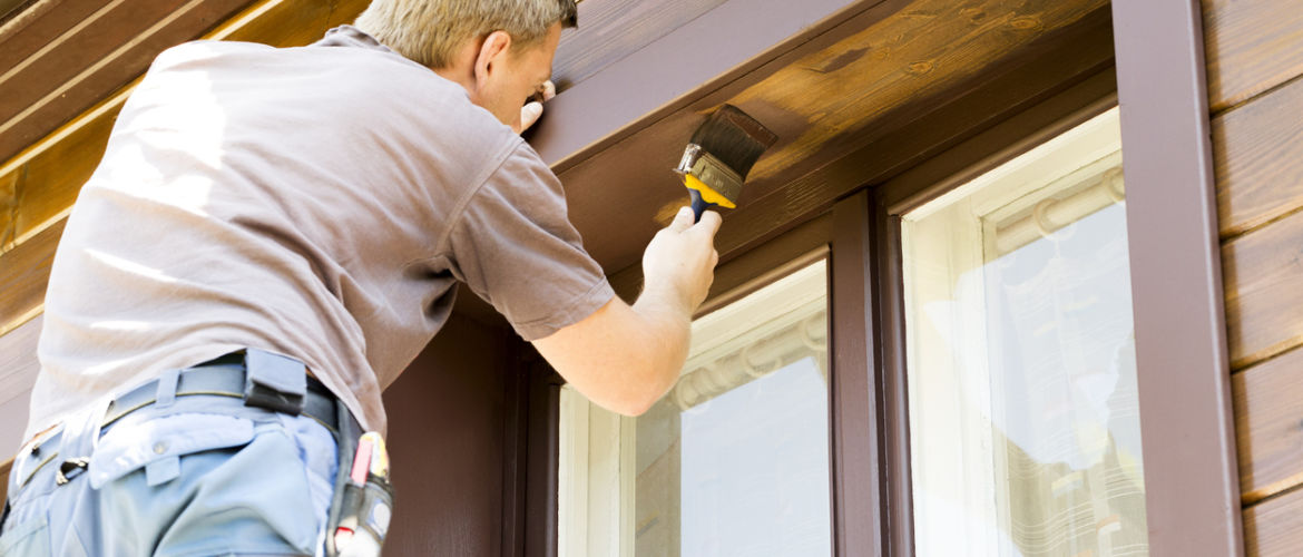 man with paintbrush painting brown wooden house exterior